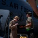 Two U.S. Air Force personnel are conducting a pre-flight or post-flight inspection beside a large military aircraft marked 'U.S. AIR FORCE' with a National Guard insignia. A woman in a flight suit is reviewing documents on a clipboard held by a second person in a t-shirt and cap. The scene appears to take place on a flight line in a sunny, arid environment.