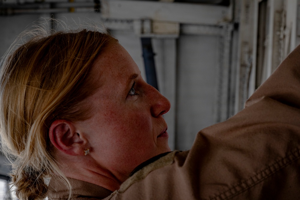 A close-up profile photograph of a woman with reddish-blonde hair and freckles, wearing what appears to be a tan/khaki military or work uniform. She is looking upward at an angle in what appears to be an industrial or military facility interior. The lighting is dramatic with shadows emphasizing her facial features.
