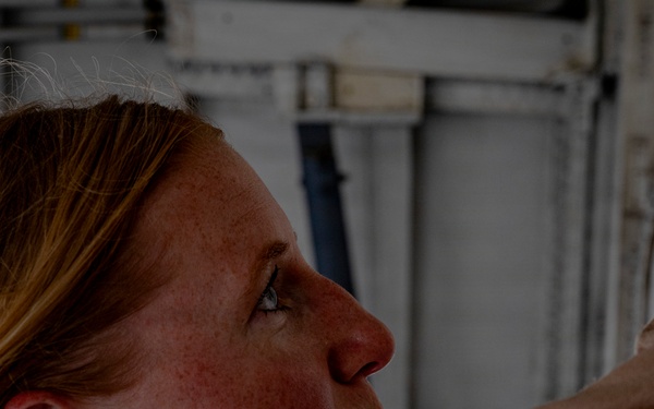 A close-up profile photograph of a woman with reddish-blonde hair and freckles, wearing what appears to be a tan/khaki military or work uniform. She is looking upward at an angle in what appears to be an industrial or military facility interior. The lighting is dramatic with shadows emphasizing her facial features.