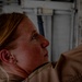 A close-up profile photograph of a woman with reddish-blonde hair and freckles, wearing what appears to be a tan/khaki military or work uniform. She is looking upward at an angle in what appears to be an industrial or military facility interior. The lighting is dramatic with shadows emphasizing her facial features.