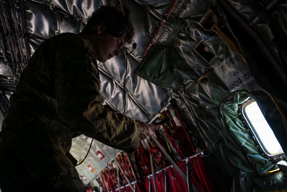 A military personnel member in camouflage uniform is working inside what appears to be a military transport aircraft, likely a C-130 or similar cargo aircraft. The individual is wearing a communications headset and appears to be inspecting or adjusting equipment near the aircraft's interior wall. The interior shows typical military aircraft features including insulated walls, safety straps, and emergency equipment markings.