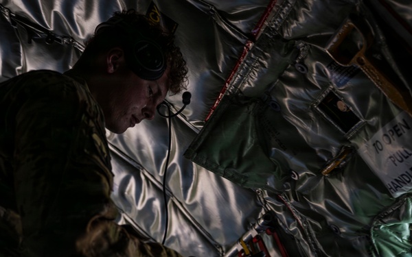A military personnel member in camouflage uniform is working inside what appears to be a military transport aircraft, likely a C-130 or similar cargo aircraft. The individual is wearing a communications headset and appears to be inspecting or adjusting equipment near the aircraft's interior wall. The interior shows typical military aircraft features including insulated walls, safety straps, and emergency equipment markings.