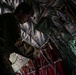 A military personnel member in camouflage uniform is working inside what appears to be a military transport aircraft, likely a C-130 or similar cargo aircraft. The individual is wearing a communications headset and appears to be inspecting or adjusting equipment near the aircraft's interior wall. The interior shows typical military aircraft features including insulated walls, safety straps, and emergency equipment markings.