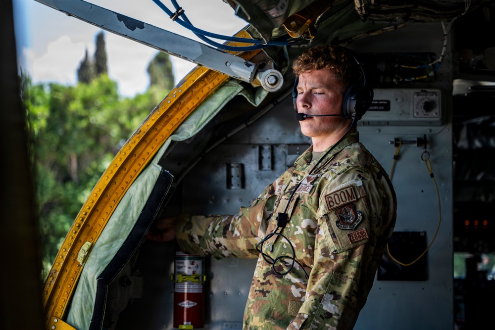 A U.S. Air Force airman wearing OCP (Operational Camouflage Pattern) uniform is stationed inside what appears to be a military aircraft, identifiable by the 'BOOM' patch indicating he is a boom operator. He is wearing a communications headset with a boom microphone and appears to be monitoring or operating equipment near an open aircraft door or hatch with yellow safety markings. The setting suggests an active aerial refueling tanker aircraft operation.
