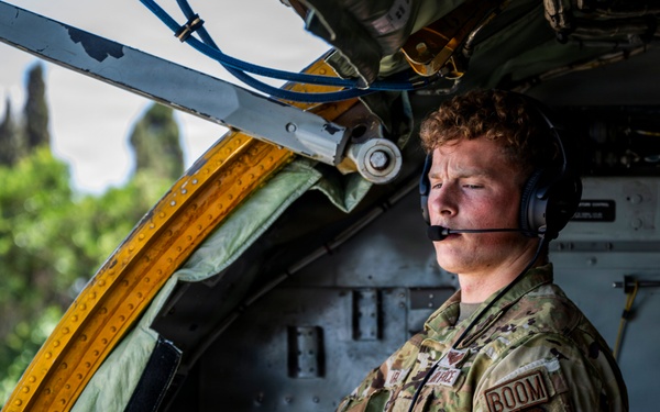 A U.S. Air Force airman wearing OCP (Operational Camouflage Pattern) uniform is stationed inside what appears to be a military aircraft, identifiable by the 'BOOM' patch indicating he is a boom operator. He is wearing a communications headset with a boom microphone and appears to be monitoring or operating equipment near an open aircraft door or hatch with yellow safety markings. The setting suggests an active aerial refueling tanker aircraft operation.