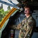A U.S. Air Force airman wearing OCP (Operational Camouflage Pattern) uniform is stationed inside what appears to be a military aircraft, identifiable by the 'BOOM' patch indicating he is a boom operator. He is wearing a communications headset with a boom microphone and appears to be monitoring or operating equipment near an open aircraft door or hatch with yellow safety markings. The setting suggests an active aerial refueling tanker aircraft operation.