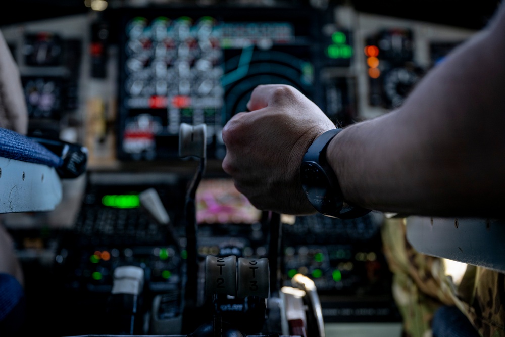 A close-up photograph taken inside a military or large transport aircraft cockpit, showing a pilot's hand gripping a control yoke or throttle assembly. The cockpit features a dense array of analog and digital instruments, switches, and illuminated indicators. The person appears to be wearing a smartwatch and military-style clothing.