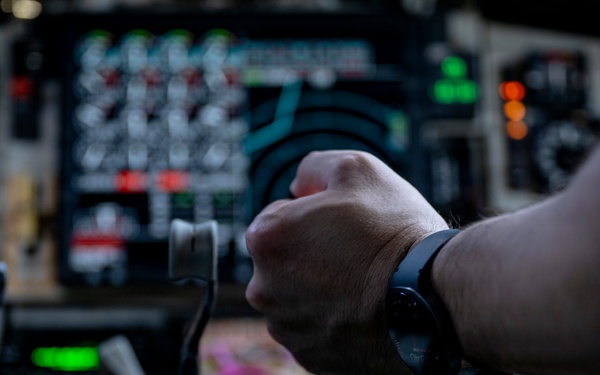 A close-up photograph taken inside a military or large transport aircraft cockpit, showing a pilot's hand gripping a control yoke or throttle assembly. The cockpit features a dense array of analog and digital instruments, switches, and illuminated indicators. The person appears to be wearing a smartwatch and military-style clothing.