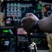 A close-up photograph taken inside a military or large transport aircraft cockpit, showing a pilot's hand gripping a control yoke or throttle assembly. The cockpit features a dense array of analog and digital instruments, switches, and illuminated indicators. The person appears to be wearing a smartwatch and military-style clothing.