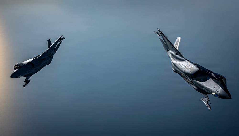 Two modern stealth fighter jets are captured in a dramatic banking maneuver against an overcast, blue-grey sky. Both aircraft appear to be fifth-generation fighters, with the left aircraft in a steep left bank and the right aircraft in a near-vertical climb or roll. The image is a high-quality aerial photograph likely taken from a chase aircraft or third aircraft flying in formation.