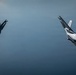 Two modern stealth fighter jets are captured in a dramatic banking maneuver against an overcast, blue-grey sky. Both aircraft appear to be fifth-generation fighters, with the left aircraft in a steep left bank and the right aircraft in a near-vertical climb or roll. The image is a high-quality aerial photograph likely taken from a chase aircraft or third aircraft flying in formation.