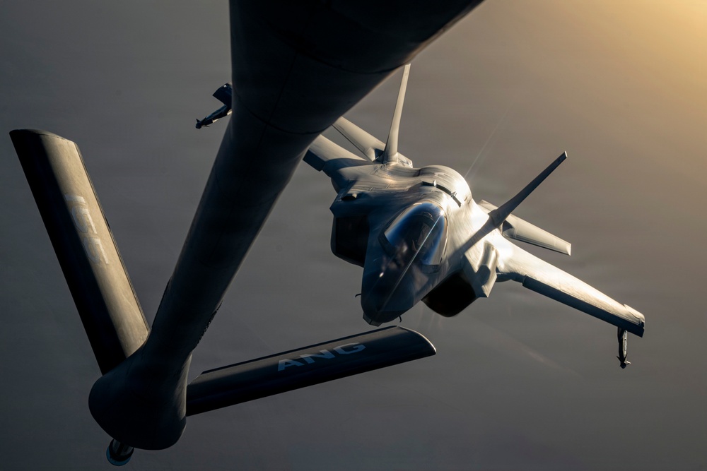 An aerial refueling operation is captured in dramatic low-light conditions, showing a fighter jet — identifiable as an F-35 — receiving fuel from a tanker aircraft. The tanker's refueling boom is visibly connected to the fighter's receptacle. The letters 'ANG' (Air National Guard) are visible on the tanker's tail section, and warm golden light from the setting or rising sun illuminates the scene from the upper right.