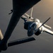 An aerial refueling operation is captured in dramatic low-light conditions, showing a fighter jet — identifiable as an F-35 — receiving fuel from a tanker aircraft. The tanker's refueling boom is visibly connected to the fighter's receptacle. The letters 'ANG' (Air National Guard) are visible on the tanker's tail section, and warm golden light from the setting or rising sun illuminates the scene from the upper right.