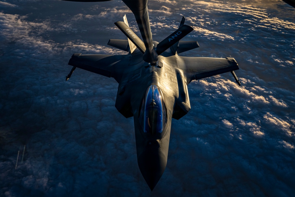 An aerial refueling operation is captured from above, showing a fighter jet — consistent with an F-35 Lightning II — receiving fuel from a tanker aircraft via a boom refueling system. The image is taken from the tanker's perspective looking down at the receiving aircraft, with a dramatic cloud layer visible far below. The letters 'ANG' (Air National Guard) are visible on the refueling boom.