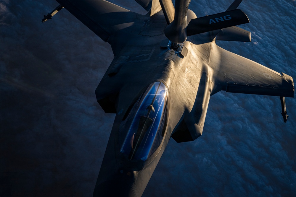 An aerial refueling operation is captured from above, showing a stealth fighter aircraft — consistent with an F-35 — receiving fuel from a tanker aircraft. The refueling boom from the tanker is visibly connected to the receiver aircraft's dorsal refueling port. The image is taken at high altitude above cloud cover, illuminated by dramatic low-angle sunlight.