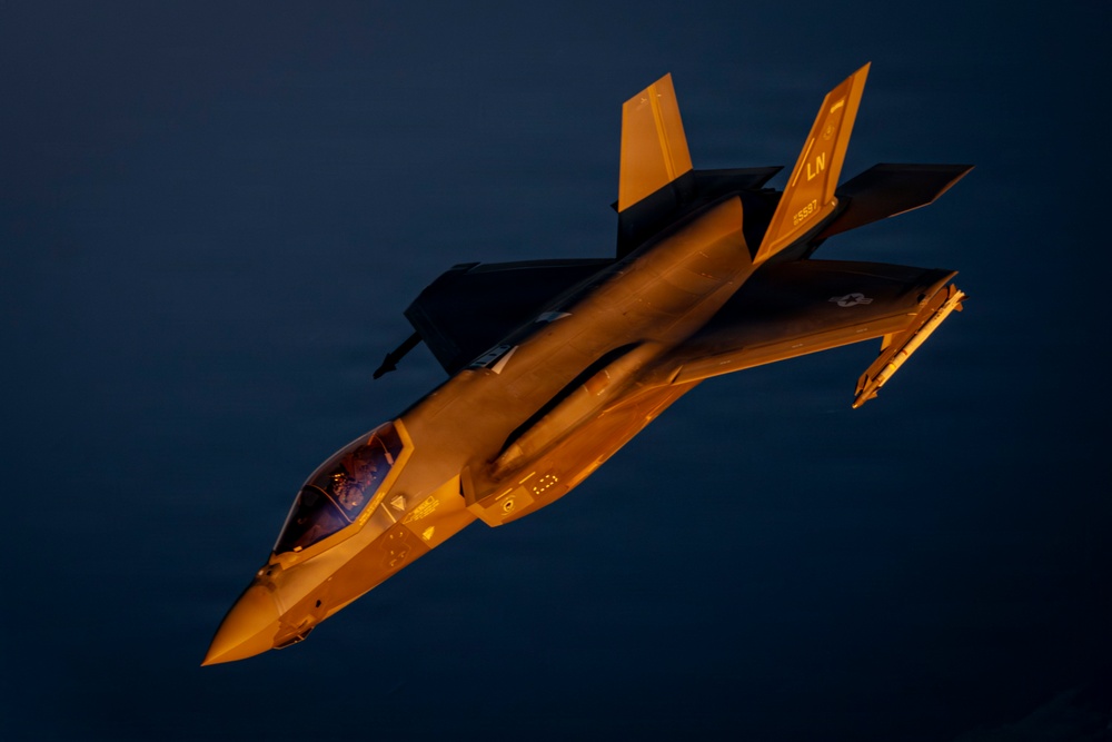 A fifth-generation stealth fighter jet, identifiable as an F-35A Lightning II, is photographed in flight during golden hour or sunset conditions. The aircraft bears the tail code 'LN', indicating assignment to Lakenheath Air Base (RAF Lakenheath, UK), home of the 48th Fighter Wing. The warm amber light dramatically illuminates the aircraft against a deep blue sky background.