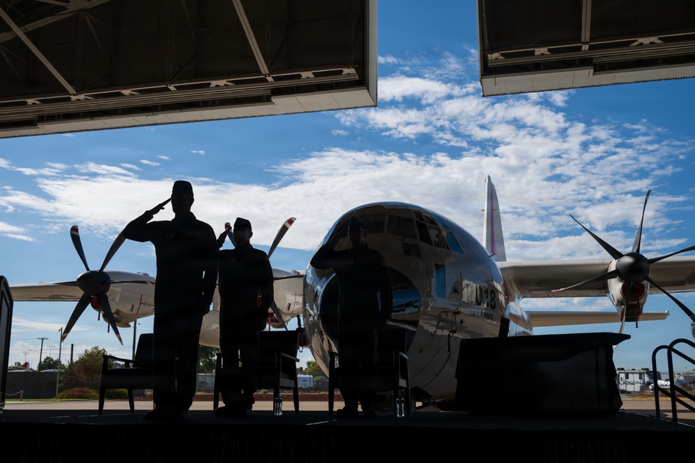 Two military personnel in silhouette are rendering a salute inside a hangar, with a large propeller-driven aircraft visible in the background against a partly cloudy sky. The scene appears to be a formal military ceremony or commemoration, likely involving the aircraft on display. The strong backlit composition creates dramatic silhouettes against the bright exterior.