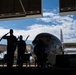 Two military personnel in silhouette are rendering a salute inside a hangar, with a large propeller-driven aircraft visible in the background against a partly cloudy sky. The scene appears to be a formal military ceremony or commemoration, likely involving the aircraft on display. The strong backlit composition creates dramatic silhouettes against the bright exterior.