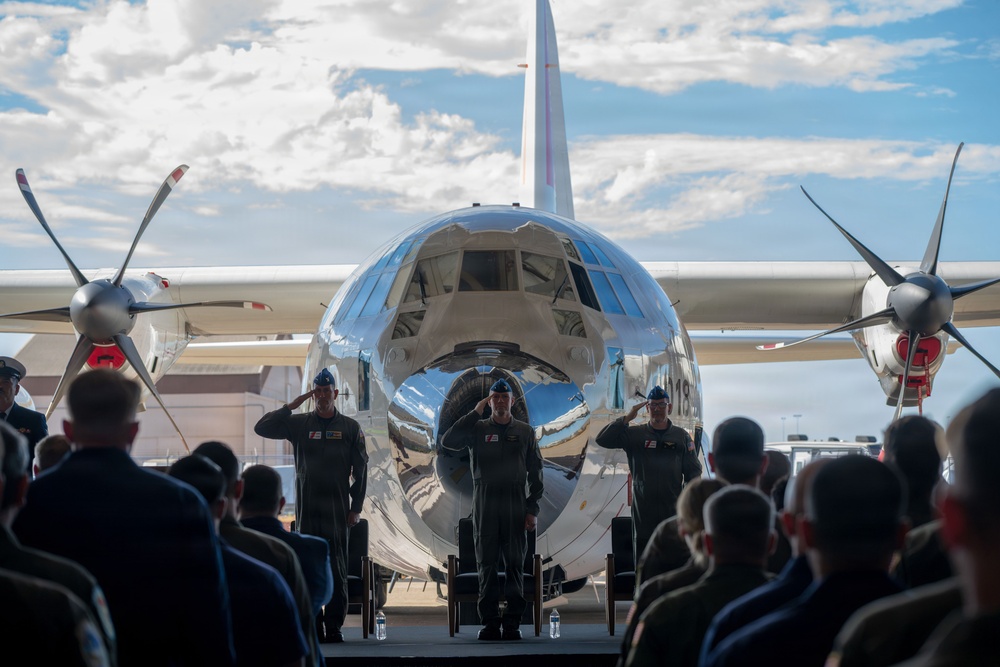A military ceremony is taking place on an airfield tarmac in front of a highly polished, mirror-finish propeller aircraft. Three uniformed military personnel are standing at attention and saluting in front of the aircraft, facing an audience of seated and standing military members. The aircraft's reflective fuselage creates a striking visual backdrop for what appears to be a formal change of command or retirement ceremony.