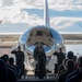 A military ceremony is taking place on an airfield tarmac in front of a highly polished, mirror-finish propeller aircraft. Three uniformed military personnel are standing at attention and saluting in front of the aircraft, facing an audience of seated and standing military members. The aircraft's reflective fuselage creates a striking visual backdrop for what appears to be a formal change of command or retirement ceremony.