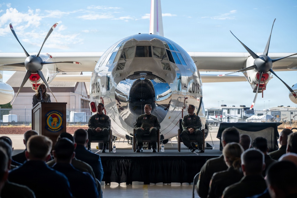 A formal military ceremony is taking place on an outdoor stage in front of a highly polished, mirror-finish turboprop aircraft. Three uniformed military personnel are seated on stage while a speaker addresses the audience from a podium bearing a military unit insignia. A large audience of uniformed personnel is seated facing the stage.