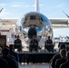 A formal military ceremony is taking place on an outdoor stage in front of a highly polished, mirror-finish turboprop aircraft. Three uniformed military personnel are seated on stage while a speaker addresses the audience from a podium bearing a military unit insignia. A large audience of uniformed personnel is seated facing the stage.