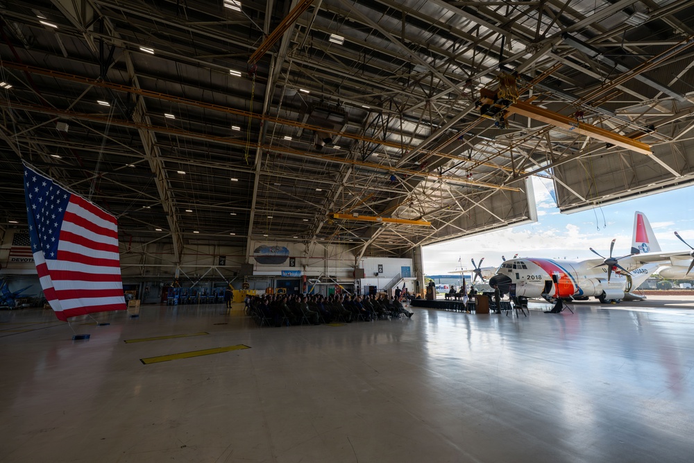 A formal military or official ceremony is taking place inside a large aviation hangar. An audience is seated in rows of chairs facing a podium and stage area, with a large U.S. flag prominently displayed on the left. A U.S. Coast Guard HC-130 aircraft (numbered 2018) is visible through the open hangar doors on the right.