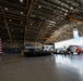 A formal military or official ceremony is taking place inside a large aviation hangar. An audience is seated in rows of chairs facing a podium and stage area, with a large U.S. flag prominently displayed on the left. A U.S. Coast Guard HC-130 aircraft (numbered 2018) is visible through the open hangar doors on the right.