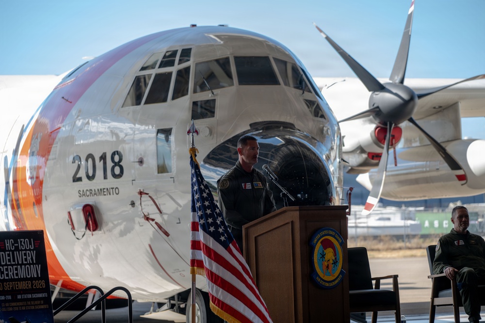 A military officer in flight suit delivers remarks at a podium during an HC-130J delivery ceremony at Air Station Sacramento, dated September 2026. The event takes place on a tarmac in front of a large Coast Guard HC-130J aircraft bearing the number '2018' and the word 'Sacramento' on its nose. An American flag and a United States Coast Guard unit seal are prominently displayed at the podium.