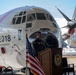 A military officer in flight suit delivers remarks at a podium during an HC-130J delivery ceremony at Air Station Sacramento, dated September 2026. The event takes place on a tarmac in front of a large Coast Guard HC-130J aircraft bearing the number '2018' and the word 'Sacramento' on its nose. An American flag and a United States Coast Guard unit seal are prominently displayed at the podium.