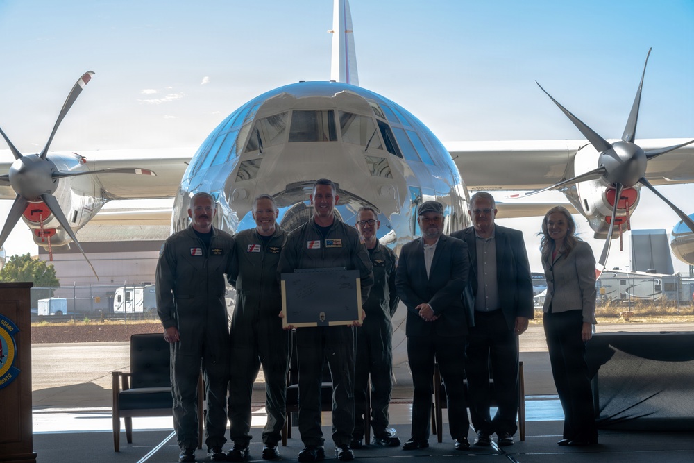 A group of seven individuals pose in front of a large, highly polished silver propeller aircraft on what appears to be an airfield tarmac. The group includes four people in military flight suits and three civilians in business attire, with one person holding a framed plaque or award. The setting suggests a formal ceremony or presentation event.