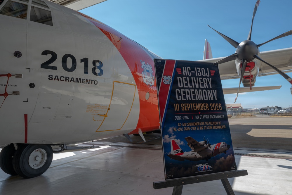 The image shows a U.S. Coast Guard HC-130J aircraft numbered 2018, assigned to Air Station Sacramento, during a delivery ceremony. A ceremonial poster on an easel in the foreground announces the 'HC-130J Delivery Ceremony' dated 10 September 2026 for CGNR-2018 at Air Station Sacramento. The aircraft displays the distinctive Coast Guard orange and white livery with the Coast Guard shield insignia.