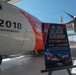 The image shows a U.S. Coast Guard HC-130J aircraft numbered 2018, assigned to Air Station Sacramento, during a delivery ceremony. A ceremonial poster on an easel in the foreground announces the 'HC-130J Delivery Ceremony' dated 10 September 2026 for CGNR-2018 at Air Station Sacramento. The aircraft displays the distinctive Coast Guard orange and white livery with the Coast Guard shield insignia.