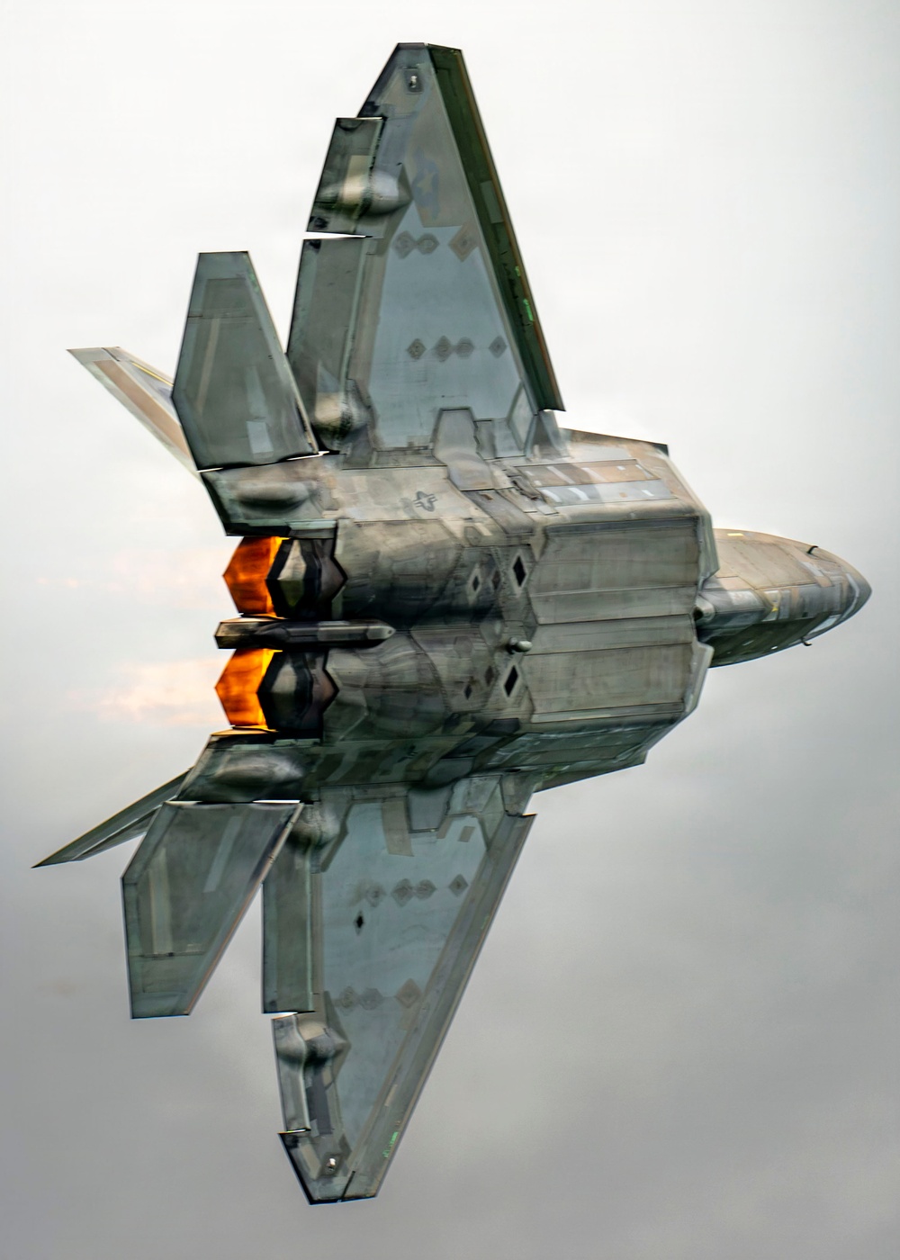 A U.S. Air Force F-22 Raptor fifth-generation stealth fighter jet is captured in a near-vertical climb maneuver, viewed from below and behind. Both afterburners are visibly ignited, producing bright orange-yellow exhaust flames. The aircraft's stealthy angular geometry, twin canted vertical stabilizers, and low-observable surface features are clearly visible.