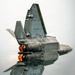 A U.S. Air Force F-22 Raptor fifth-generation stealth fighter jet is captured in a near-vertical climb maneuver, viewed from below and behind. Both afterburners are visibly ignited, producing bright orange-yellow exhaust flames. The aircraft's stealthy angular geometry, twin canted vertical stabilizers, and low-observable surface features are clearly visible.