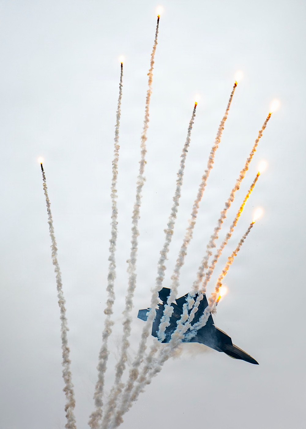 A military stealth fighter jet, consistent with an F-22 Raptor, is captured mid-maneuver deploying a large spread of flares in an overcast sky. The aircraft appears to be in a steep banking or diving attitude while simultaneously releasing multiple pyrotechnic countermeasure flares that fan outward in a dramatic arc. This appears to be an airshow demonstration showcasing the aircraft's flare dispensing capability.
