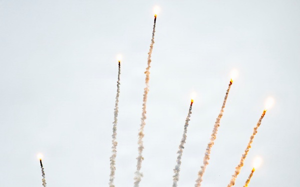 A military stealth fighter jet, consistent with an F-22 Raptor, is captured mid-maneuver deploying a large spread of flares in an overcast sky. The aircraft appears to be in a steep banking or diving attitude while simultaneously releasing multiple pyrotechnic countermeasure flares that fan outward in a dramatic arc. This appears to be an airshow demonstration showcasing the aircraft's flare dispensing capability.