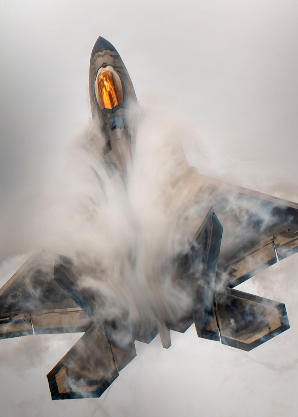 An overhead view of what appears to be an F-22 Raptor stealth fighter jet performing a high-performance maneuver, generating a dramatic vapor cone or condensation cloud around its fuselage. The aircraft's canopy is visible with a distinctive amber/gold tint, and the angular stealth geometry of the airframe is clearly visible through the swirling white condensation. The image captures a Prandtl-Glauert condensation phenomenon caused by rapid pressure changes during the maneuver.