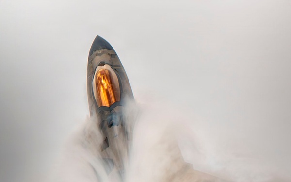 An overhead view of what appears to be an F-22 Raptor stealth fighter jet performing a high-performance maneuver, generating a dramatic vapor cone or condensation cloud around its fuselage. The aircraft's canopy is visible with a distinctive amber/gold tint, and the angular stealth geometry of the airframe is clearly visible through the swirling white condensation. The image captures a Prandtl-Glauert condensation phenomenon caused by rapid pressure changes during the maneuver.