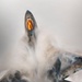 An overhead view of what appears to be an F-22 Raptor stealth fighter jet performing a high-performance maneuver, generating a dramatic vapor cone or condensation cloud around its fuselage. The aircraft's canopy is visible with a distinctive amber/gold tint, and the angular stealth geometry of the airframe is clearly visible through the swirling white condensation. The image captures a Prandtl-Glauert condensation phenomenon caused by rapid pressure changes during the maneuver.