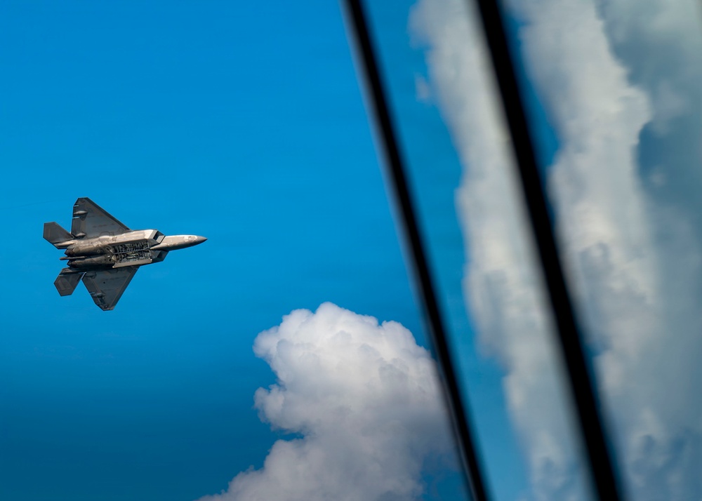 A fifth-generation stealth fighter jet, identifiable as an F-22 Raptor, is captured in flight against a vivid blue sky with cumulus clouds. The aircraft's weapons bay doors appear to be open, revealing internal payload compartments. The right portion of the image shows a reflective glass surface, likely from another aircraft, mirroring the clouds and sky.