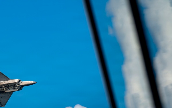 A fifth-generation stealth fighter jet, identifiable as an F-22 Raptor, is captured in flight against a vivid blue sky with cumulus clouds. The aircraft's weapons bay doors appear to be open, revealing internal payload compartments. The right portion of the image shows a reflective glass surface, likely from another aircraft, mirroring the clouds and sky.