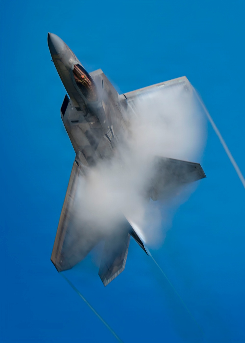 A fifth-generation stealth fighter jet, consistent with an F-22 Raptor, is captured in a near-vertical climb against a clear blue sky. The aircraft is generating a dramatic vapor cone or condensation cloud around its fuselage and wings, indicative of high-speed, high-G maneuvering. The image appears to have been taken during an airshow or aerial demonstration.