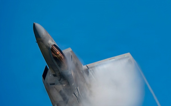 A fifth-generation stealth fighter jet, consistent with an F-22 Raptor, is captured in a near-vertical climb against a clear blue sky. The aircraft is generating a dramatic vapor cone or condensation cloud around its fuselage and wings, indicative of high-speed, high-G maneuvering. The image appears to have been taken during an airshow or aerial demonstration.