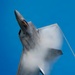 A fifth-generation stealth fighter jet, consistent with an F-22 Raptor, is captured in a near-vertical climb against a clear blue sky. The aircraft is generating a dramatic vapor cone or condensation cloud around its fuselage and wings, indicative of high-speed, high-G maneuvering. The image appears to have been taken during an airshow or aerial demonstration.
