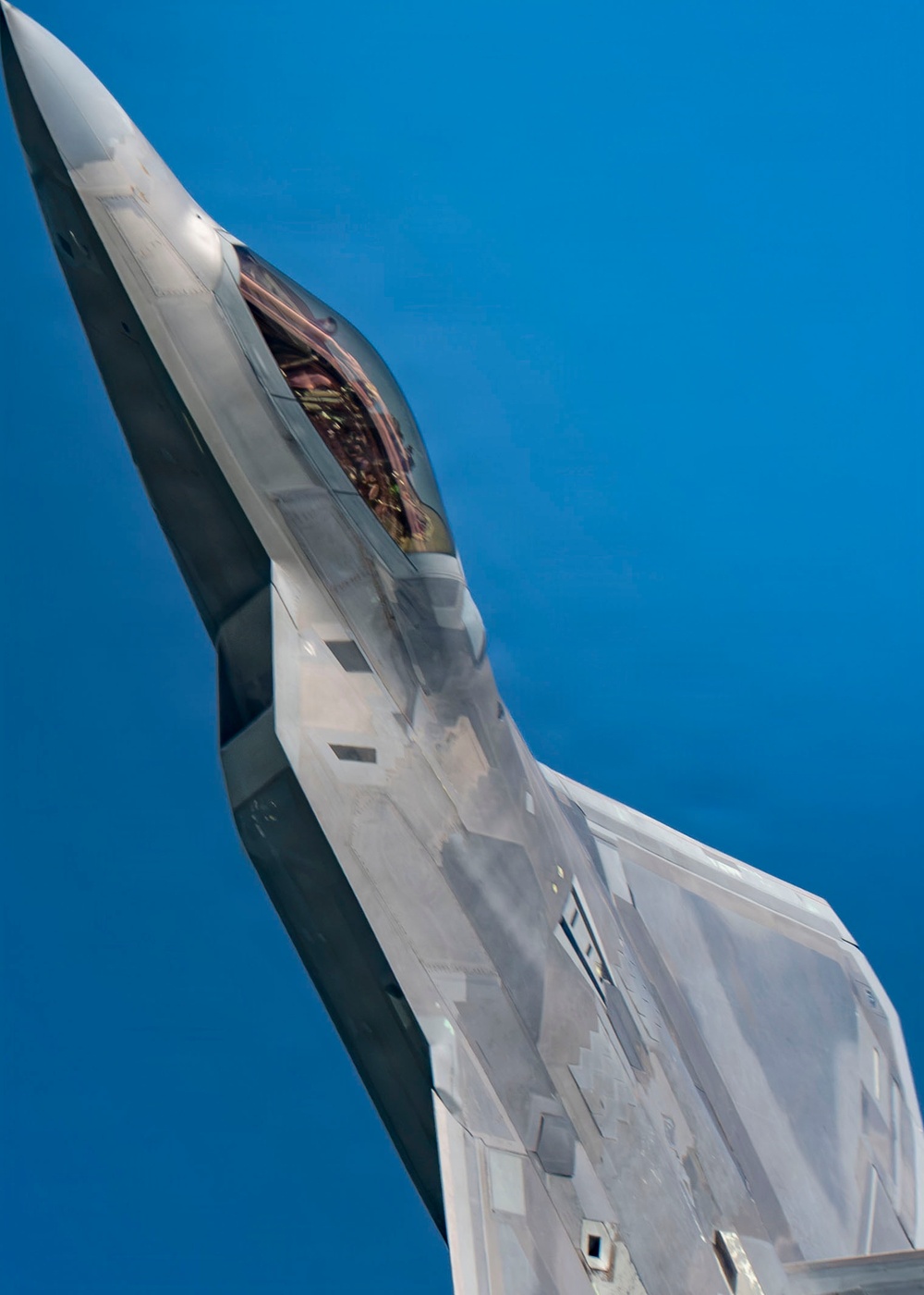 A close-up aerial photograph of what appears to be an F-22 Raptor stealth fighter jet in flight against a clear blue sky. The aircraft is captured at a steep banking angle, revealing the open cockpit canopy with a pilot visible inside. The image showcases the aircraft's distinctive angular stealth geometry and gray composite surface panels.