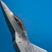 A close-up aerial photograph of what appears to be an F-22 Raptor stealth fighter jet in flight against a clear blue sky. The aircraft is captured at a steep banking angle, revealing the open cockpit canopy with a pilot visible inside. The image showcases the aircraft's distinctive angular stealth geometry and gray composite surface panels.