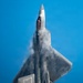 A fifth-generation stealth fighter aircraft, identifiable as an F-22 Raptor, is photographed from directly below in a near-vertical climb maneuver against a clear blue sky. Condensation vapor is visibly forming over the fuselage and wing surfaces due to rapid pressure changes during the high-performance maneuver. The image captures exceptional detail of the aircraft's underside, including its stealth-optimized angular geometry and open weapon bay doors.