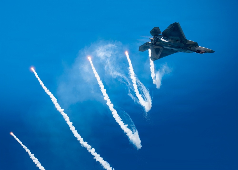 A stealth fighter jet, consistent with the F-22 Raptor design, is captured in flight against a vivid blue sky while deploying multiple flares. The flares trail bright white smoke plumes downward in a dramatic fan pattern. This appears to be an airshow demonstration or a countermeasure deployment exercise.