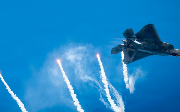 A stealth fighter jet, consistent with the F-22 Raptor design, is captured in flight against a vivid blue sky while deploying multiple flares. The flares trail bright white smoke plumes downward in a dramatic fan pattern. This appears to be an airshow demonstration or a countermeasure deployment exercise.
